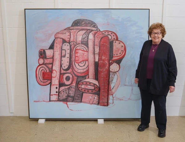 Musa Mayer stands beside a large abstract painting by Philip Guston featuring red and pink biomorphic shapes against a pale blue background.