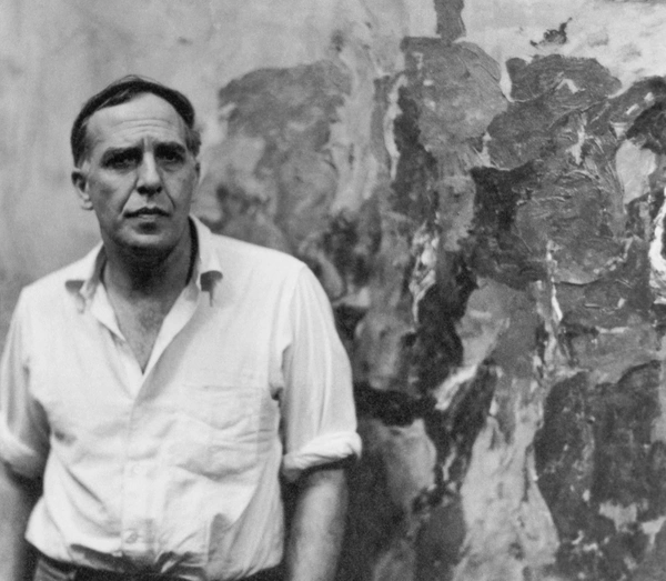 Philip Guston in a dark sweater sits in a studio, holding a cigarette, with abstract artwork hanging on the wall behind him.