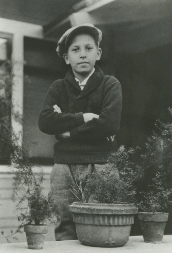 A black-and-white photograph of Philip Guston at age 10. Standing behind potted plants, arms crossed. He wears a flat cap, a dark sweater, and light-colored trousers.