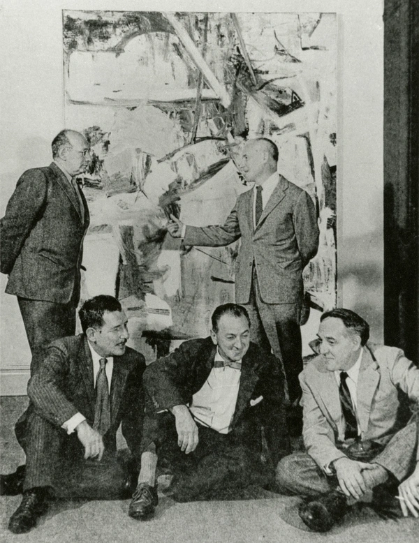 Black-and-white photograph of five men in suits gathered in front of a large abstract painting; three sit on the floor while two stand behind them, one gesturing toward the artwork.