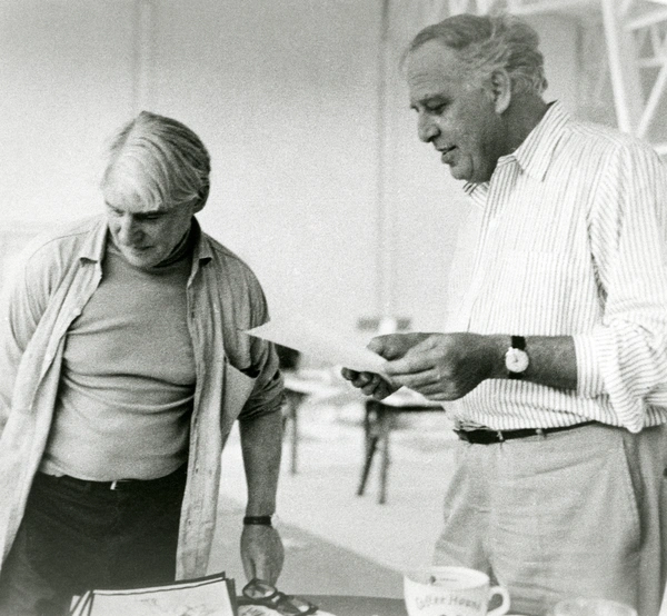 Black-and-white photograph of two older men standing indoors, looking down at papers on a table; one holds a sheet while the other leans over nearby documents and a mug.