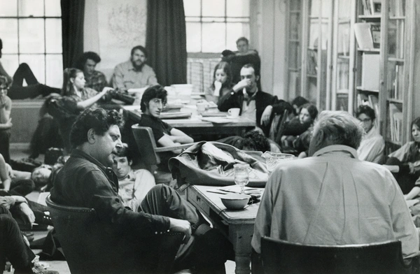 A black-and-white photo of a classroom or studio filled with students listening intently to two men, Leland Bell and Philip Guston, seated at a central table.