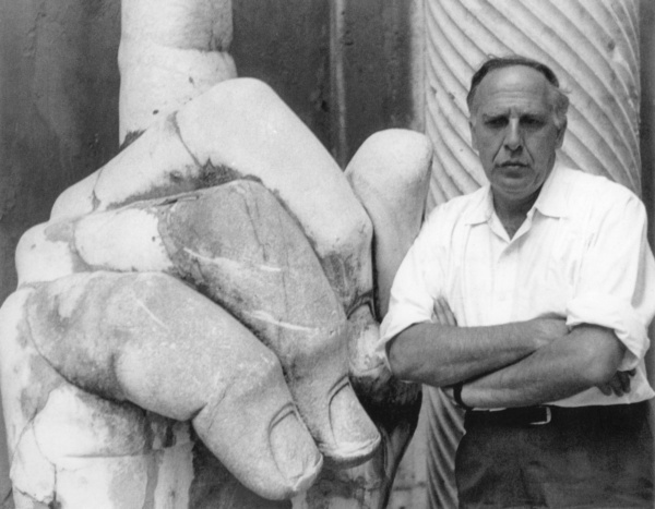 Man with arms crossed, Philip Guston, standing beside a giant carved stone hand.