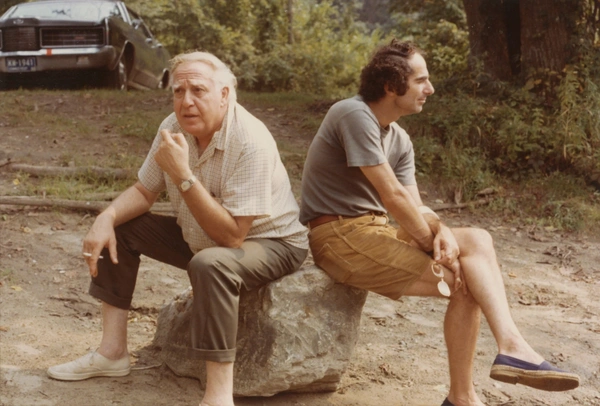 Philip Guston and Philip Roth sit back-to-back on a large rock in a wooded outdoor setting, one holding a cigarette and the other a pair of glasses.