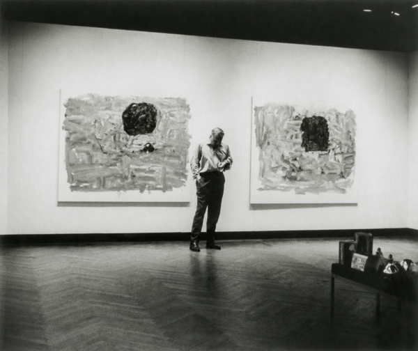 Black-and-white photo of a man, Philip Guston, standing in a gallery between two large abstract paintings, each with a dark central shape, under spotlights on a polished wood floor.