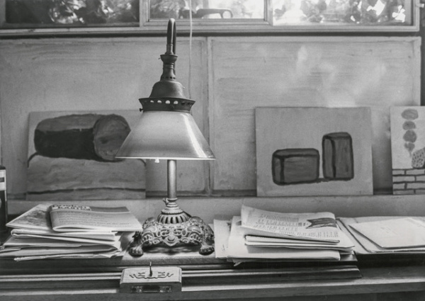 Black-and-white photo of a desk lamp surrounded by piles of papers, with Philip Guston paintings of blocky shapes propped up behind it.