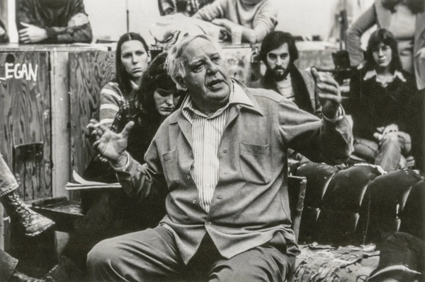 Black-and-white photo of an older man, Philip Guston,  seated among an audience, gesturing with both hands as he speaks while several people watch attentively.