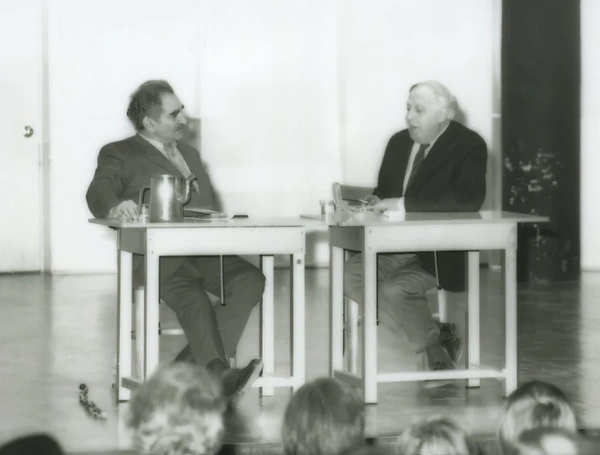Black-and-white photo of a public dialogue between two men, Harold Rosenberg and Philip Guston, at separate desks.