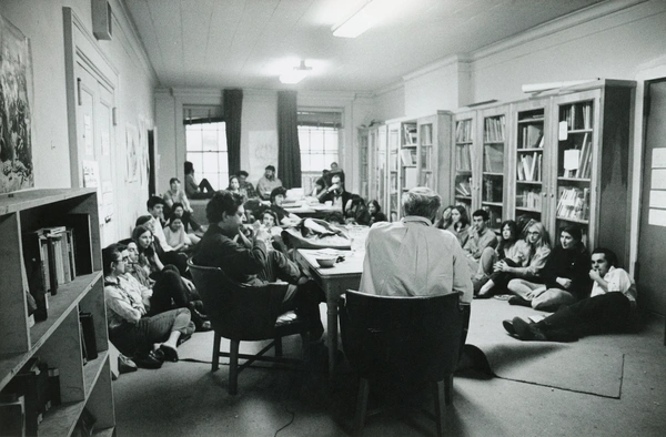 A black-and-white photo of a group of students sitting on the floor and in chairs, attentively listening to two men (Philip Guston and Leland Bell) leading a discussion at a central table in a room lined with bookshelves and posters.